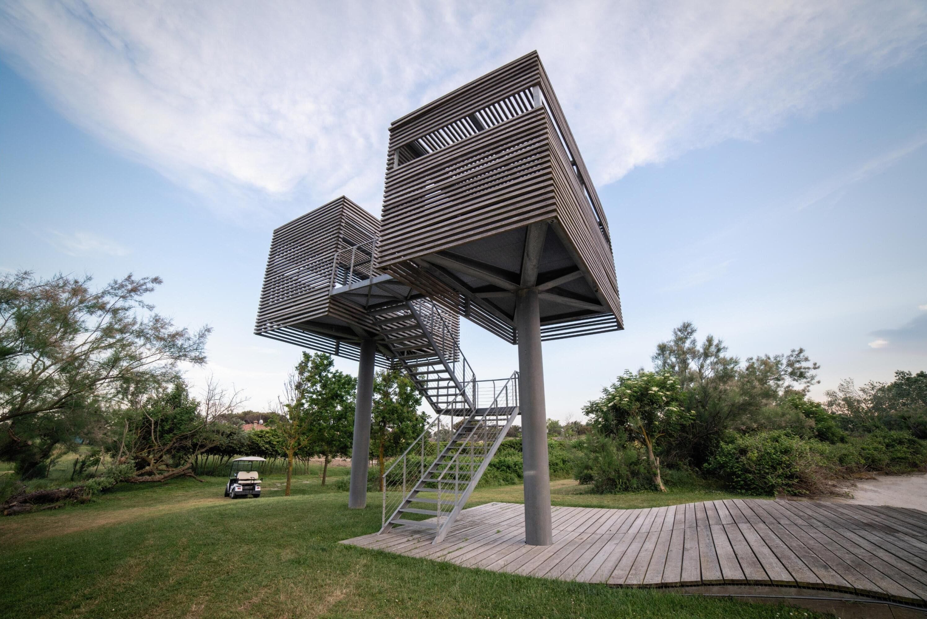 A modern outdoor structure with a wooden deck and metal railings, surrounded by greenery.