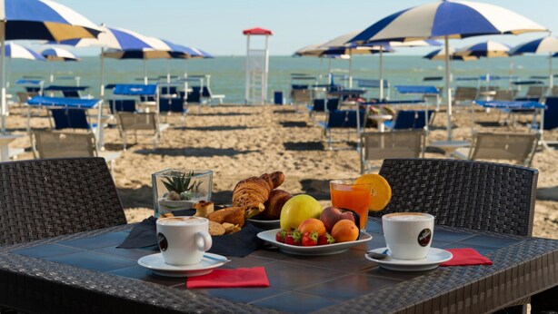 Tavolo per la colazione in spiaggia con croissant, frutta e caffè, con vista sulla spiaggia, ombrelloni e lettini.