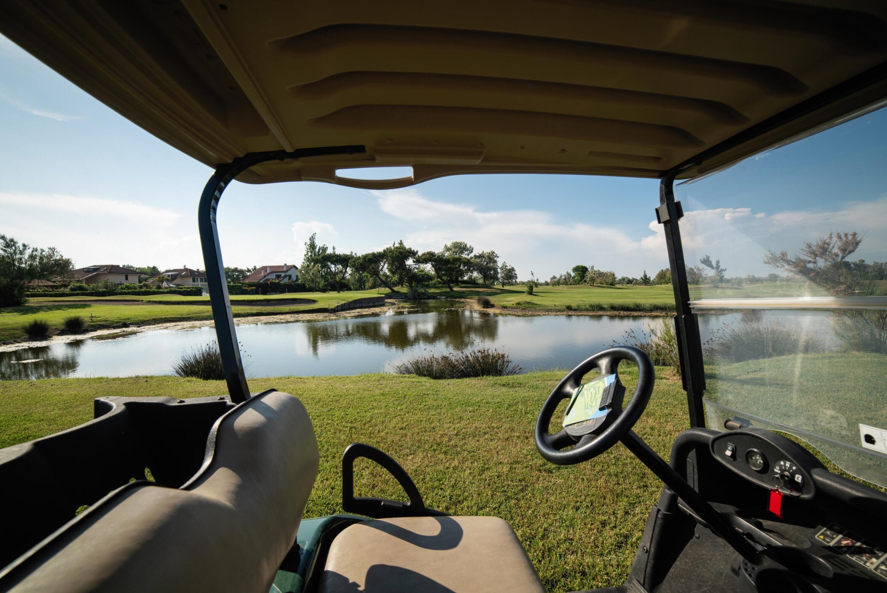 Het interieur van een golfkarretje, met stuur en dashboard, met uitzicht op een vijver en groen landschap.