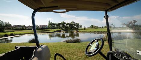 The interior of a golf cart with a steering wheel and dashboard, overlooking a pond and green landscape.