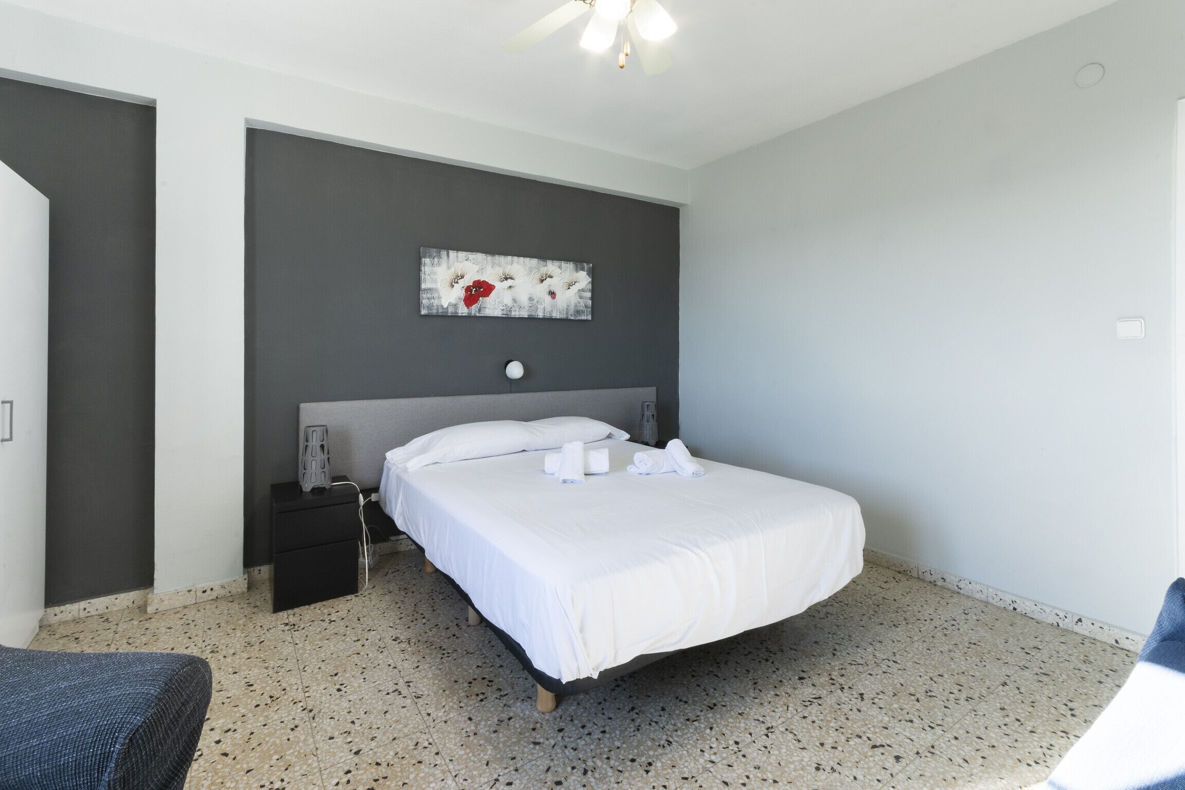 Una habitación de hotel con una cama, una mesita de noche, una obra de arte en la pared y un ventilador de techo.