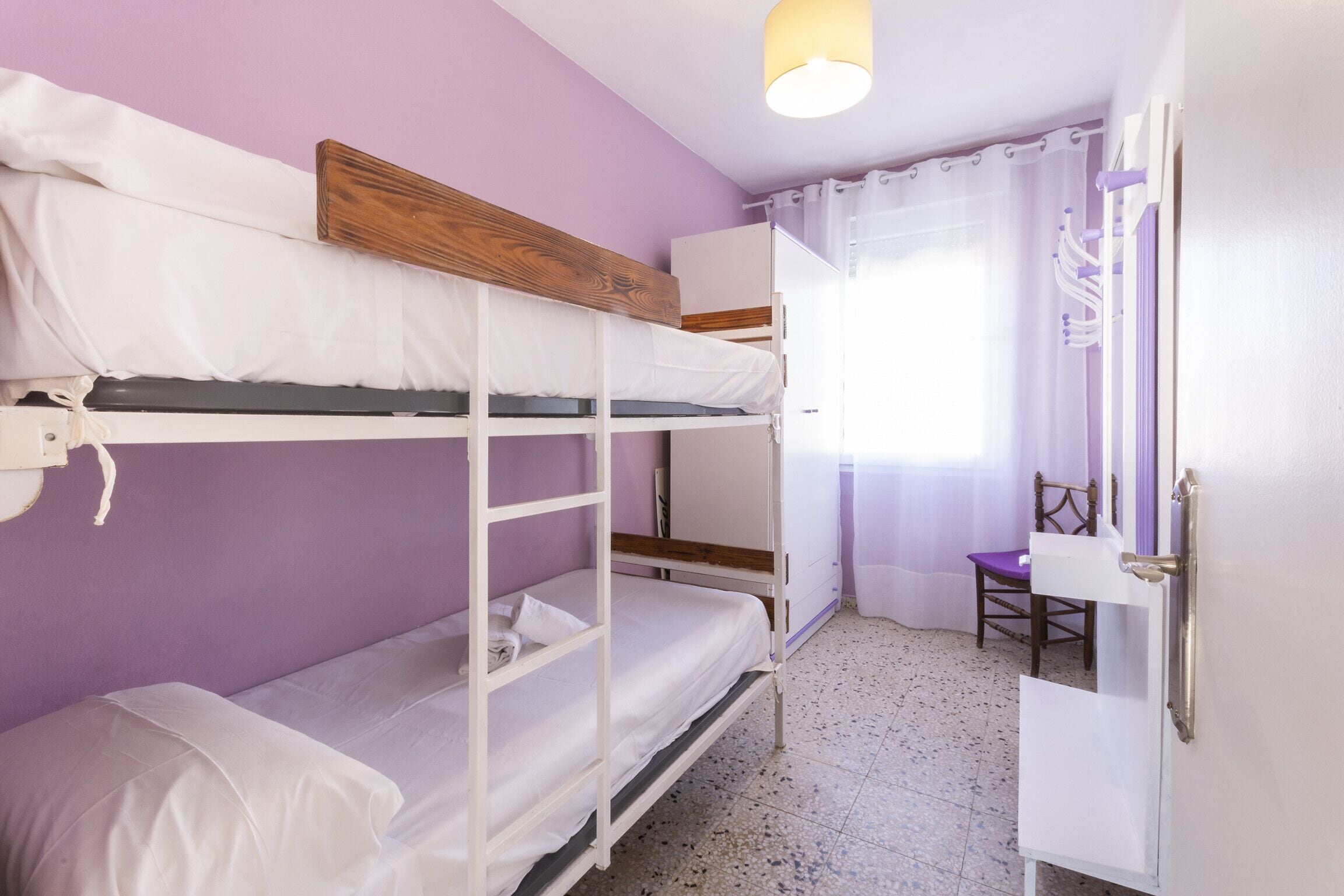 A bunk bed room with a wooden headboard, a white wardrobe, and a chair near a window with curtains.