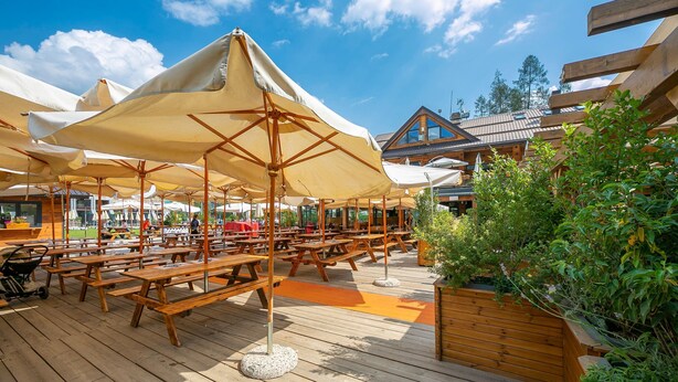 An outdoor wooden deck with picnic tables and large umbrellas, surrounded by greenery and a building with a sloped roof.