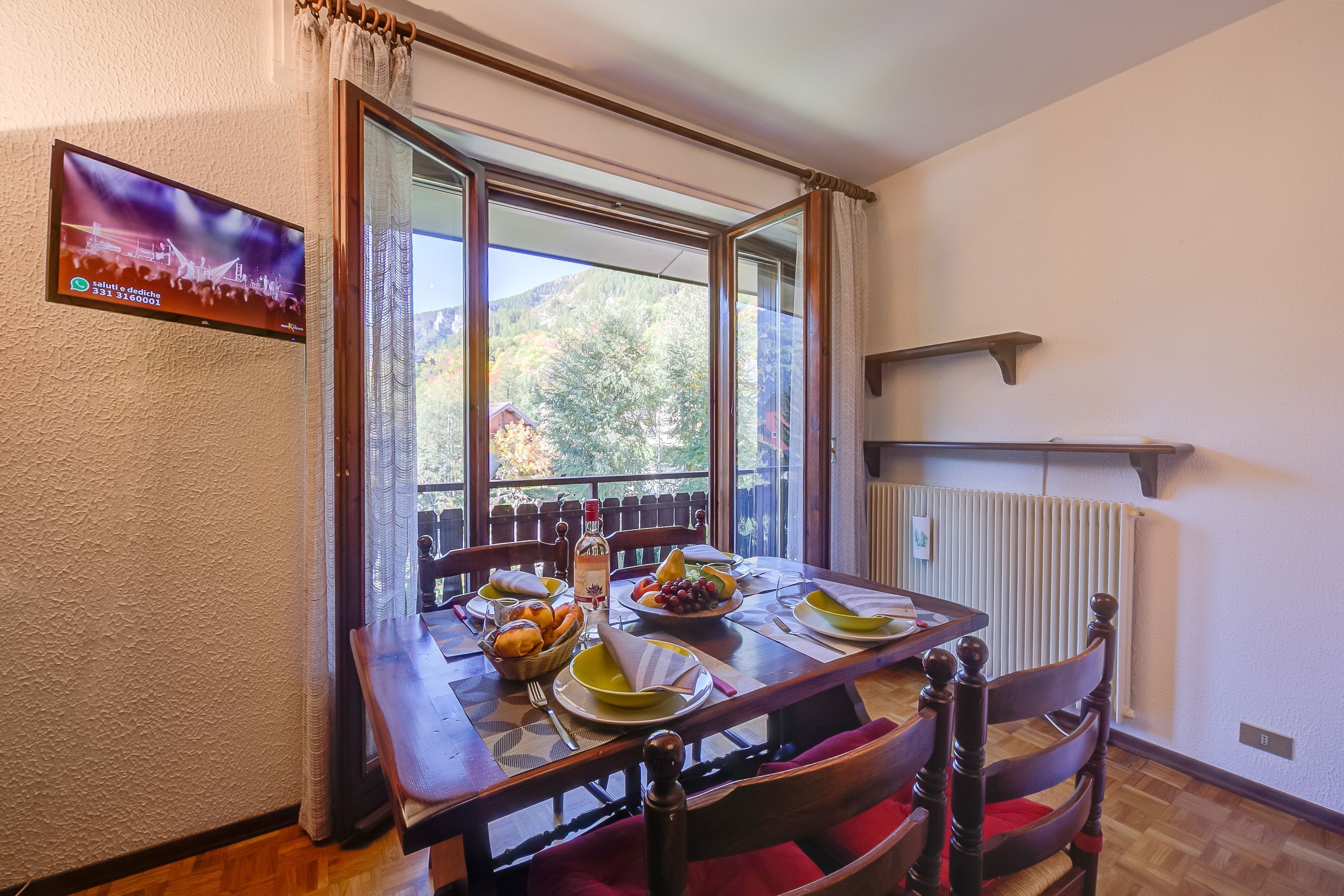A dining area with a table set for two, a TV mounted on the wall, and a view of the outdoors through a large window.