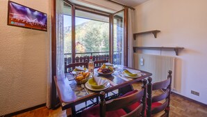 A dining area with a table set for two, a TV mounted on the wall, and a view of the outdoors through a large window. - Il Nido di Elda few Steps From Skiing (Bardonecchia)