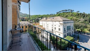 A balcony with a table and chairs, overlooking a street with parked cars and a large white building with arched windows. - Central Flat in Camogli (Camogli)