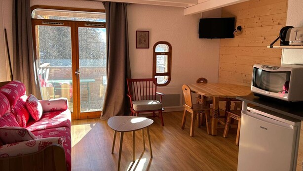 A cozy living room with a sofa, a small round table, and a TV mounted on the wall. - Renovated large studio for 4, center station, ski-in/ski-out (Les Orres)