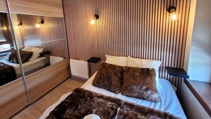 Un dormitorio moderno con un panel de pared de listones de madera, un espejo grande, una cama con ropa de cama blanca y un plaid de piel sintética, y dos mesitas de noche con lámparas.