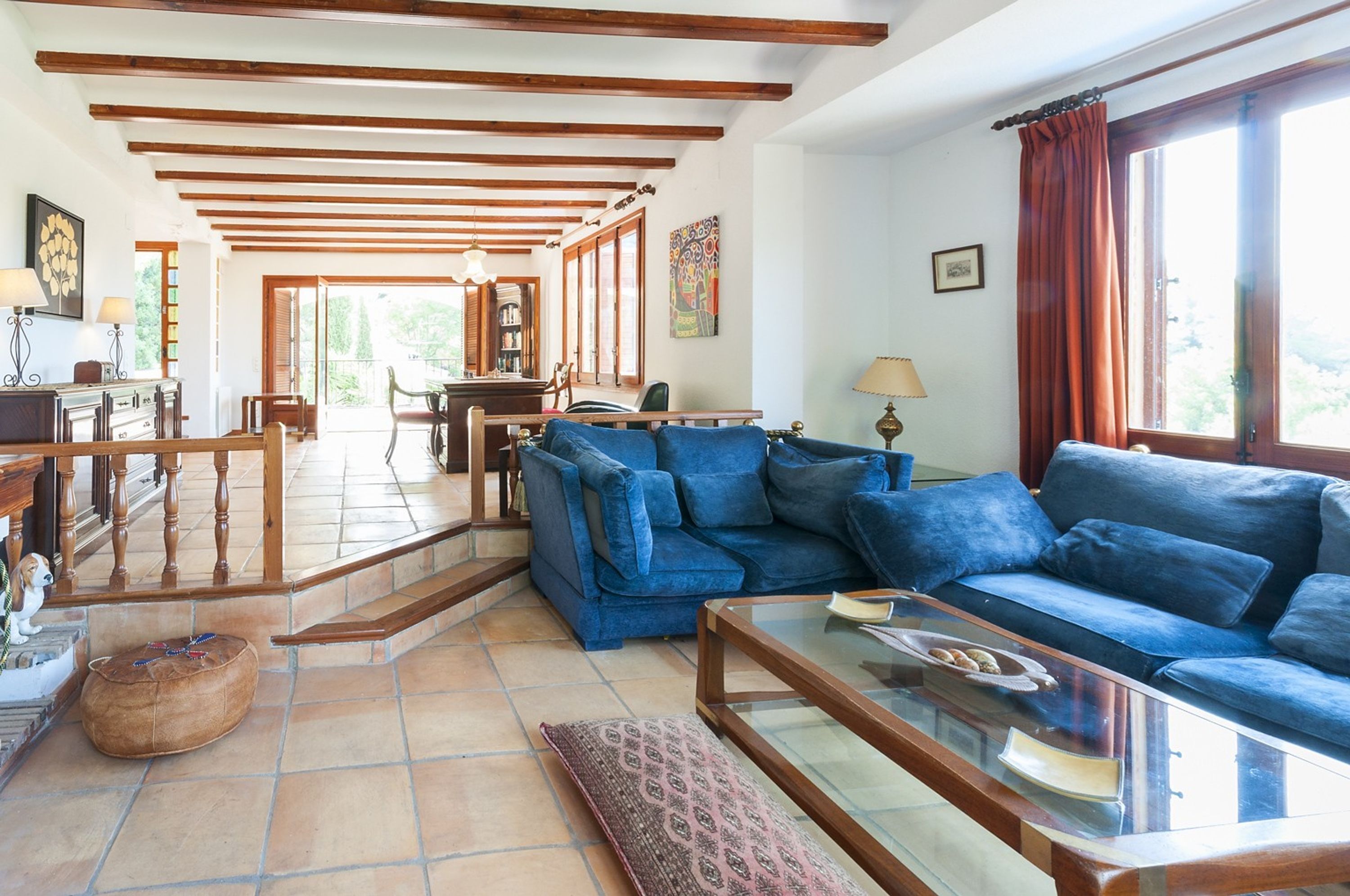 A spacious living room with a blue sofa, wooden beams on the ceiling, a glass coffee table, and a view of the outdoors.