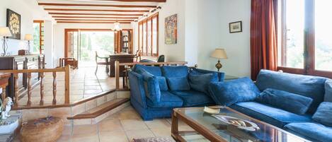 A spacious living room with a blue sofa, wooden beams on the ceiling, a glass coffee table, and a view of the outdoors.