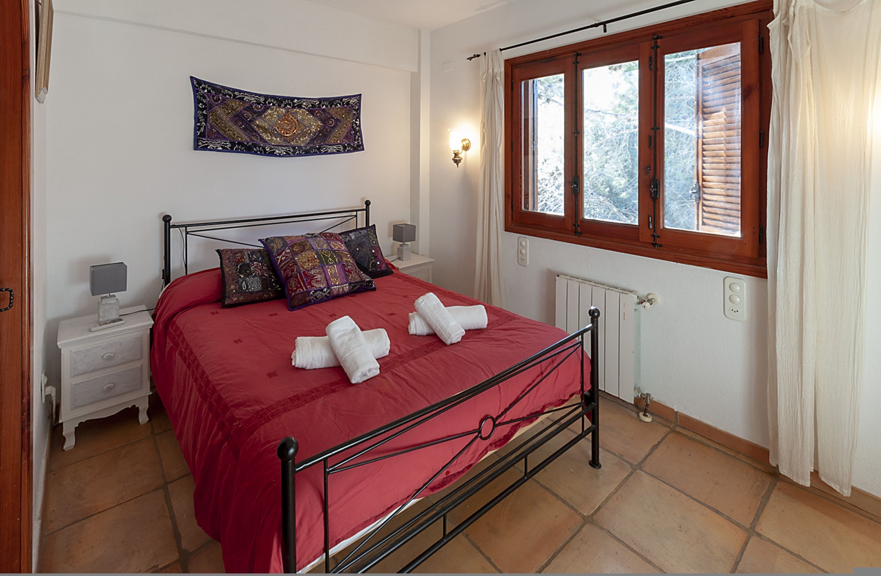 Ein Schlafzimmer mit einem Metallbettgestell, einer roten Bettdecke, zwei Nachttischen mit Lampen, einem Fenster mit Holzjalousien und einem Wandteppich.