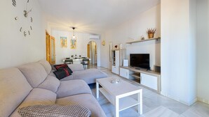 A modern living room with a grey sofa, a coffee table, and a TV. There is a kitchen area in the background with a dining table and chairs. - La Casa De Bebita in Fuengirola (Fuengirola)