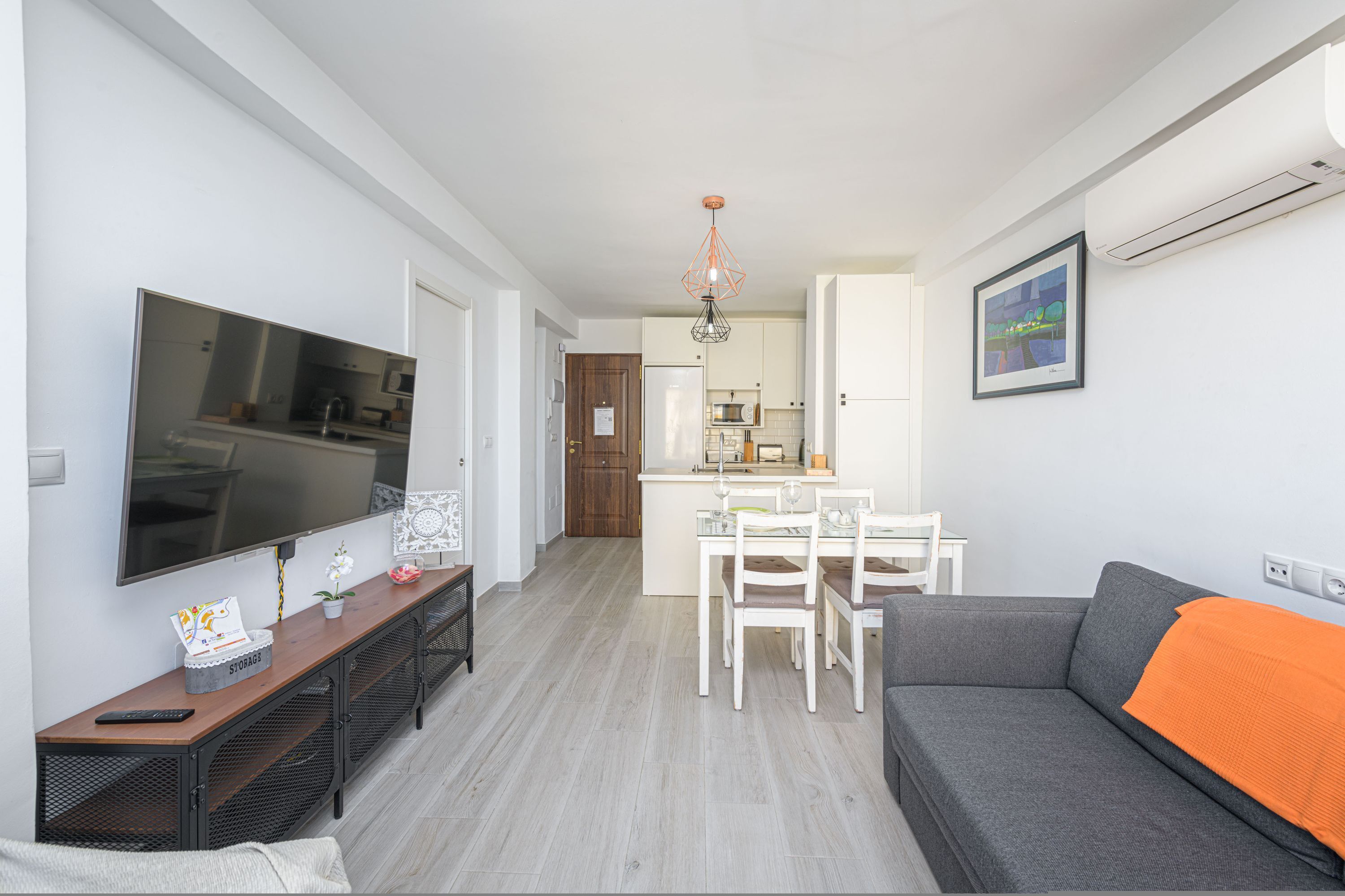 A modern living room with a grey sofa, a TV, a dining area with a white table and chairs, and a kitchen in the background.