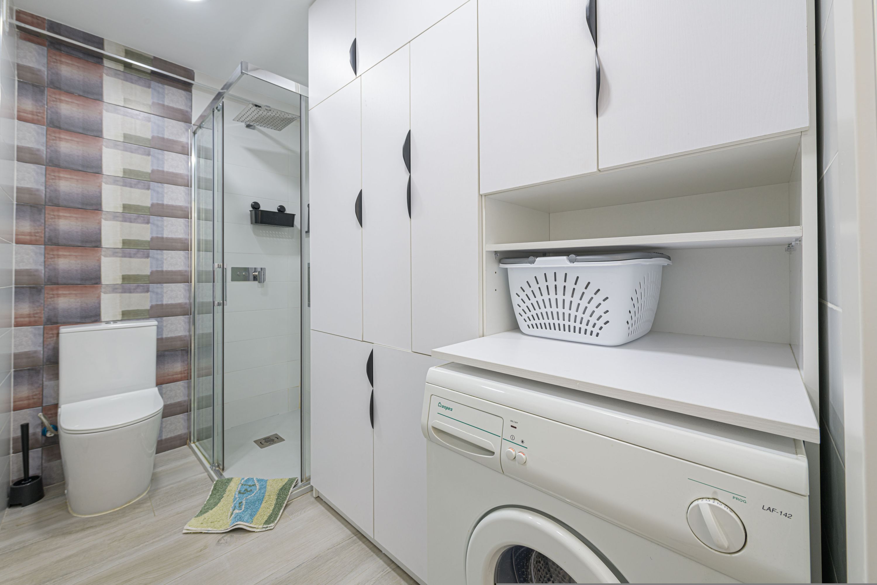 Un baño moderno con cabina de ducha de vidrio, lavadora y cesto de ropa en un estante.