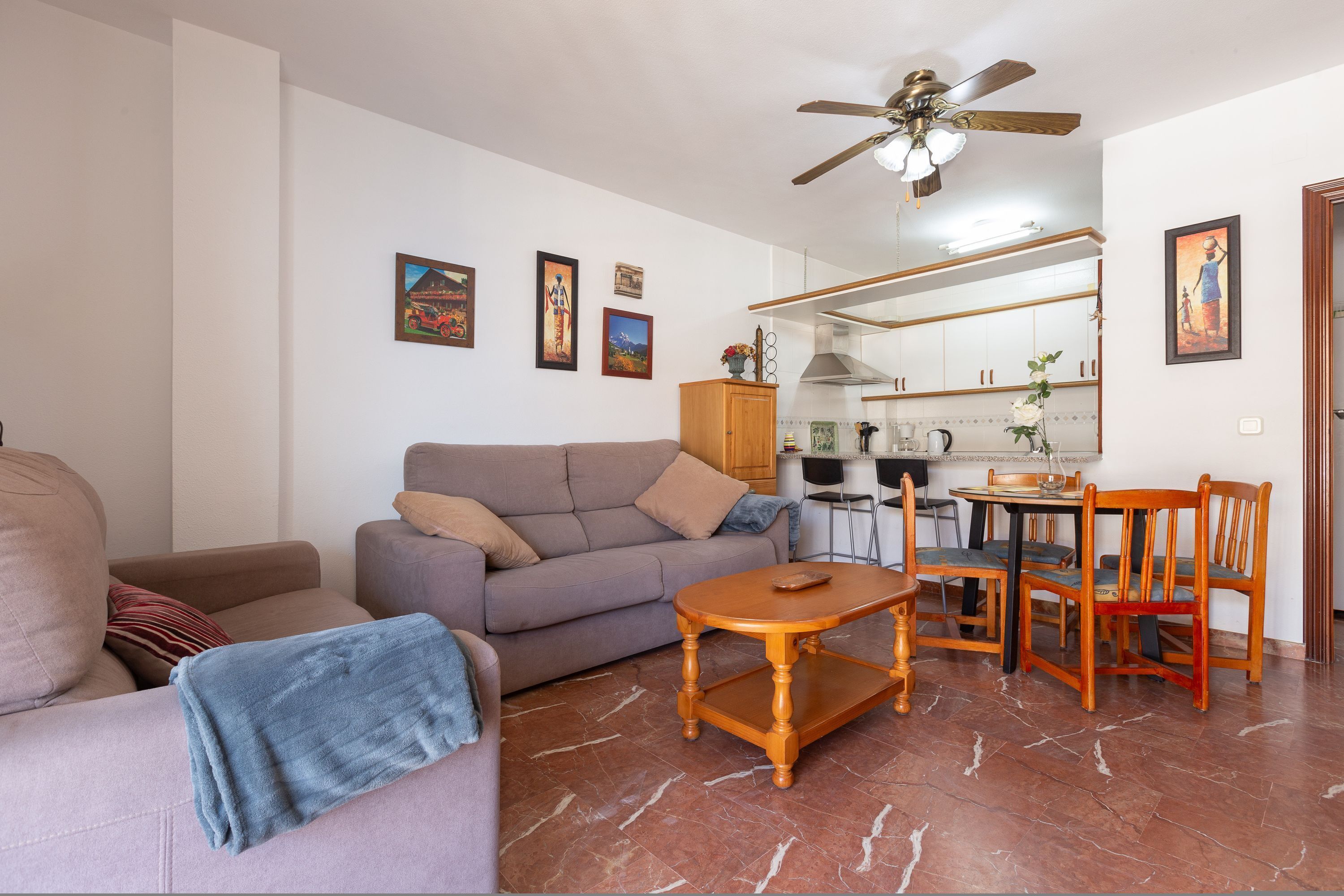 A living room with a sofa, a coffee table, and a dining area with chairs and a table. There is a ceiling fan and kitchen cabinets in the background.