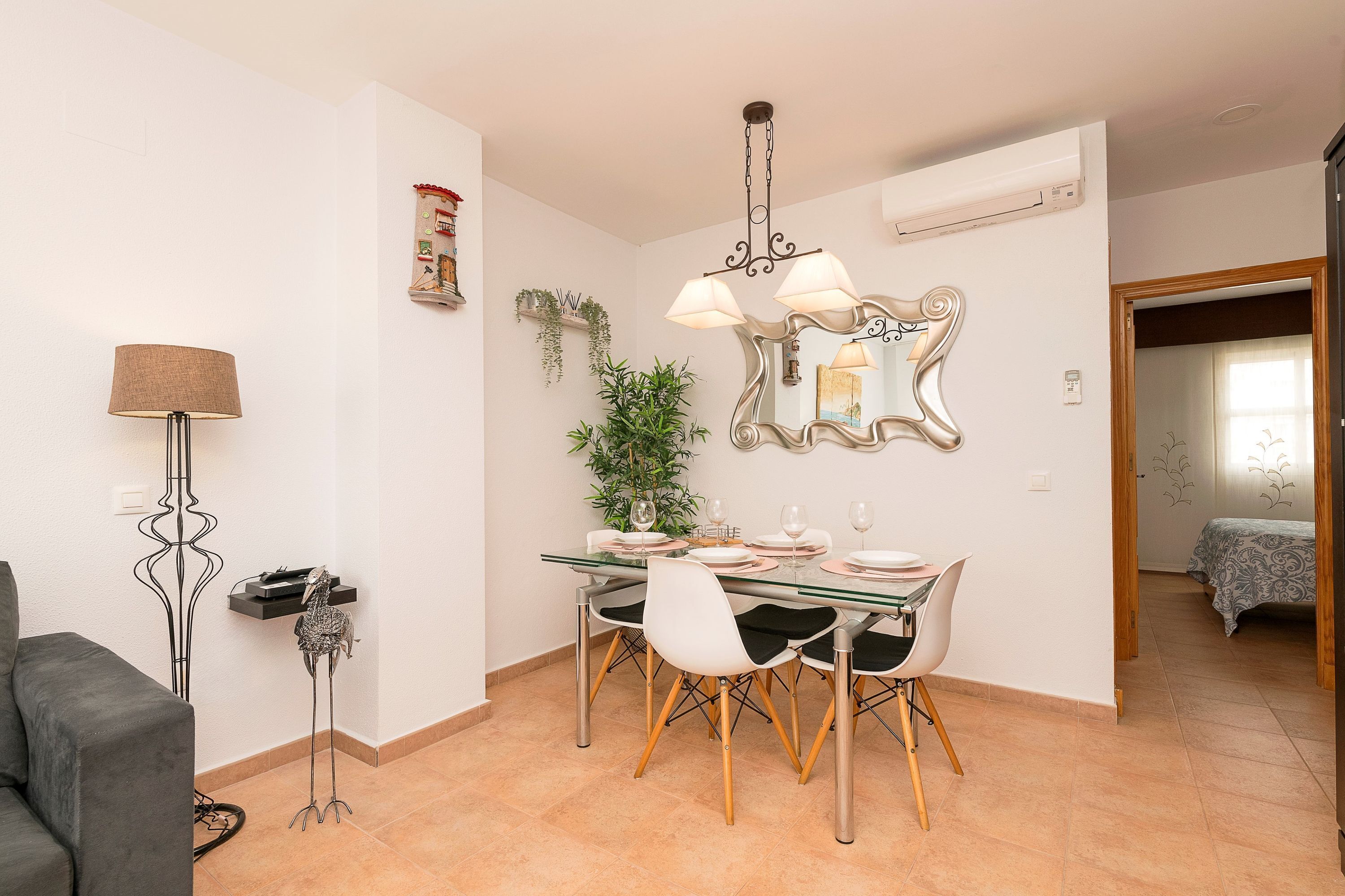 A dining area with a glass table, white chairs, and a chandelier. There is a plant on the wall and a mirror above the table.
