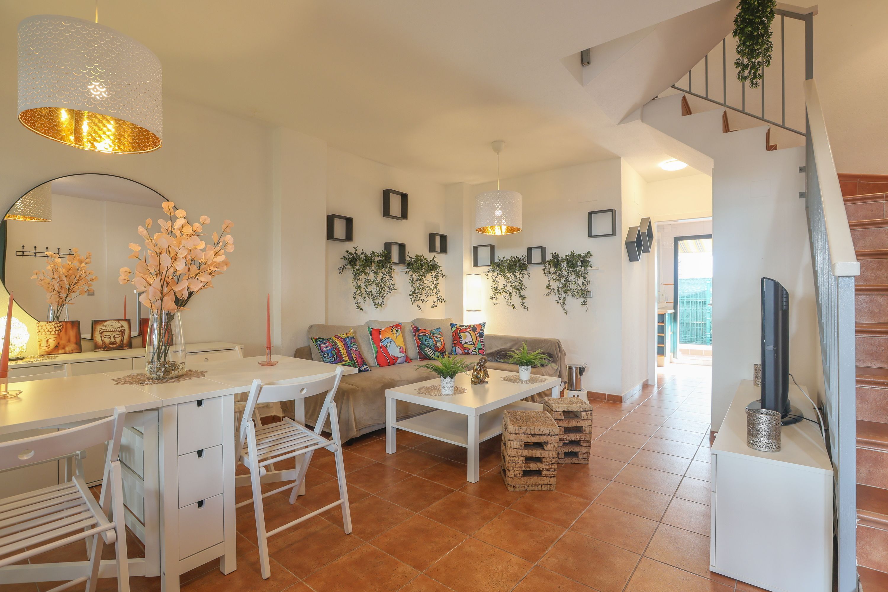 Un salon moderne avec un canapé blanc, une table à manger et des chaises. Des plantes en pot sont disposées sur les murs, et un escalier se trouve au fond de la pièce.
