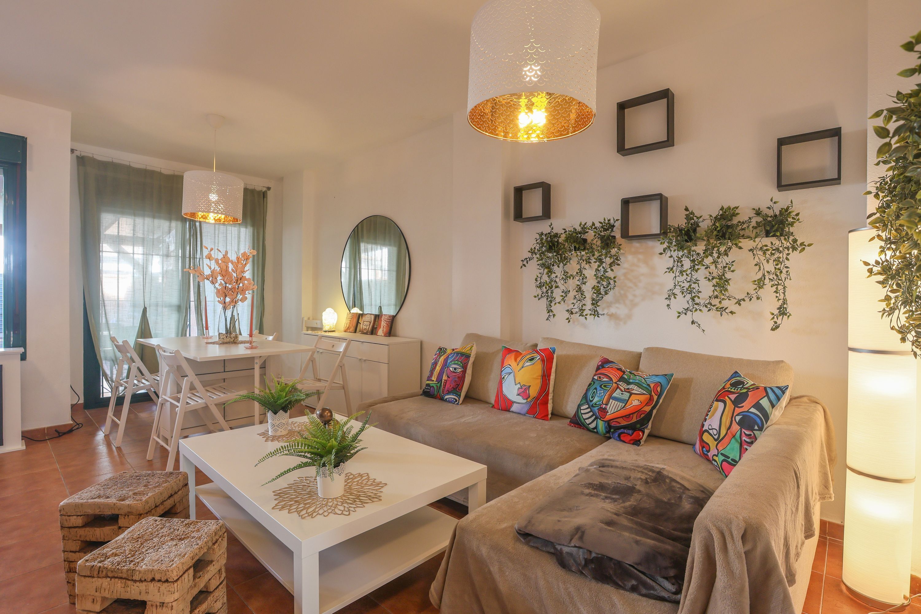 A living room with a sofa, coffee table, and chairs. There is a dining area with a table and chairs. The room has a large mirror, framed pictures, and plants on the walls.