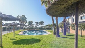 A swimming pool surrounded by grass, palm trees, and thatched umbrellas. - Magallanes in Nuevo Portil (Cartaya)