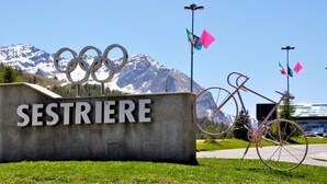 Une pancarte ornée des anneaux olympiques et du mot « SESTRIERE », avec une chaîne de montagnes en arrière-plan.