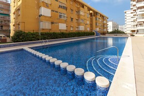 A swimming pool with blue mosaic tiles, a series of steps leading into the water, and a yellow building in the background.