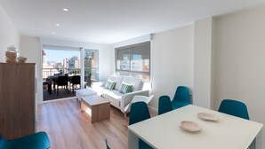 A modern living room with a white sofa, teal chairs, a dining table with decorative plates, and a view of the city through large windows. - Torres Del Mar in Grau i Platja (Gandia)