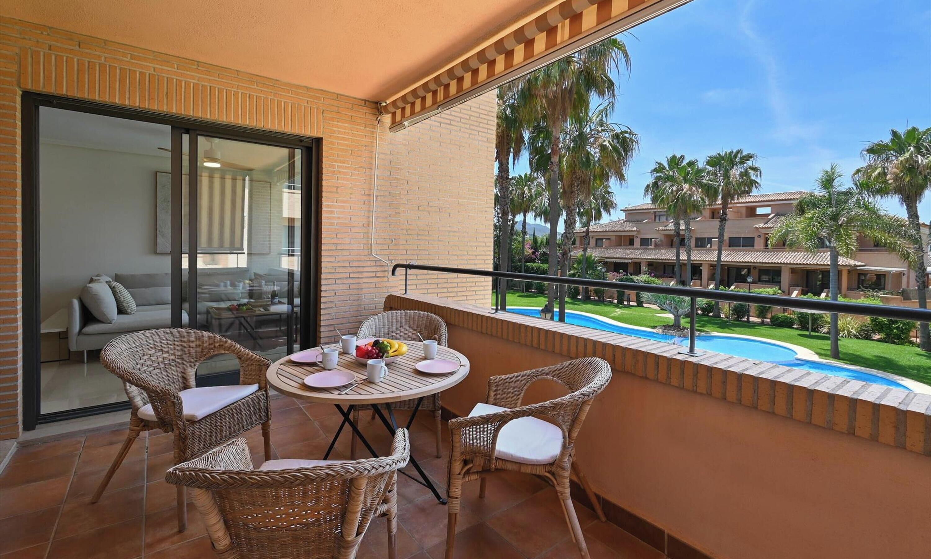 A balcony with wicker furniture, a glass door leading to a room, and a view of a pool and palm trees.