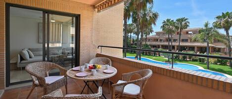 A balcony with wicker furniture, a glass door leading to a room, and a view of a pool and palm trees.