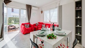 A modern living room with a red sofa, a dining table set with plates, glasses, and a bottle. There is a window with curtains, a clock on the wall, and a balcony visible through the glass door. - Serafina Beach House (San Bartolomeo al Mare)