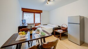 A compact room with a bed, a refrigerator, a dining table with chairs, a window with curtains, and a small table with a basket. - Studio Genzianella Ski In-ski Out Mt 100 (Sestriere)