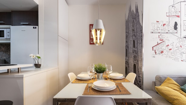 Een moderne eetruimte met een witte tafel voor zes personen, een decoratief kunstwerk aan de muur, en een keuken op de achtergrond.