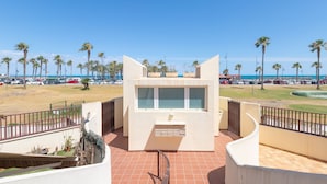 Een modern gebouw met een centrale glazen gevel, omringd door een gebogen balkon met metalen leuningen, met uitzicht op een strand met palmbomen en geparkeerde auto's.