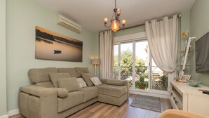 Ein Wohnzimmer mit einem beigefarbenen Ecksofa, einem Kronleuchter, einem Sonnenuntergangsbild und Blick auf Grünpflanzen durch große Fenster.