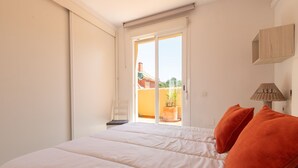 A bedroom with a bed, two orange pillows, a nightstand, a lamp, and a sliding glass door leading to an outdoor area. - Monteazul in Marbella (Marbella)