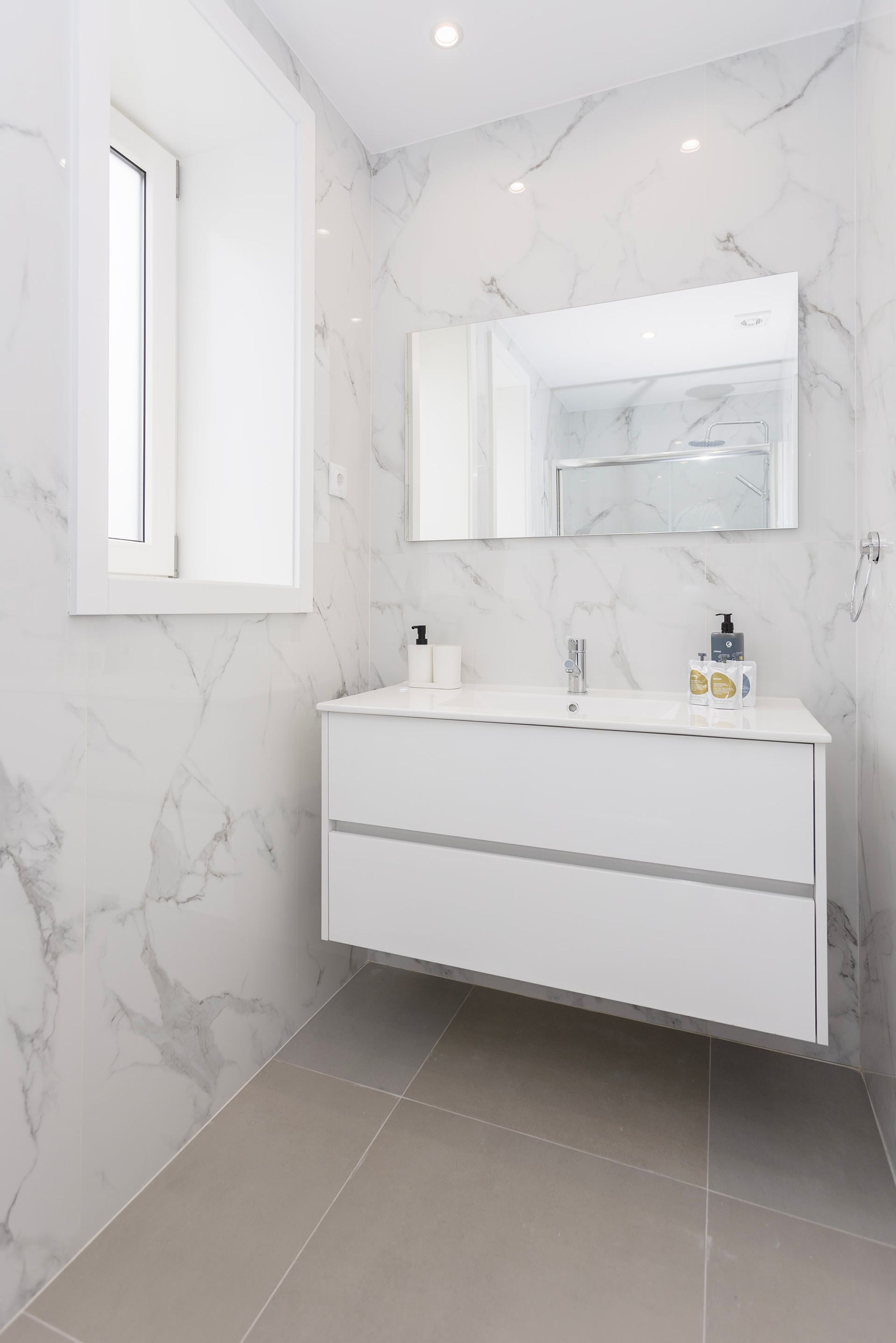 A bathroom with a marble wall, a white vanity, a mirror, and a window.
