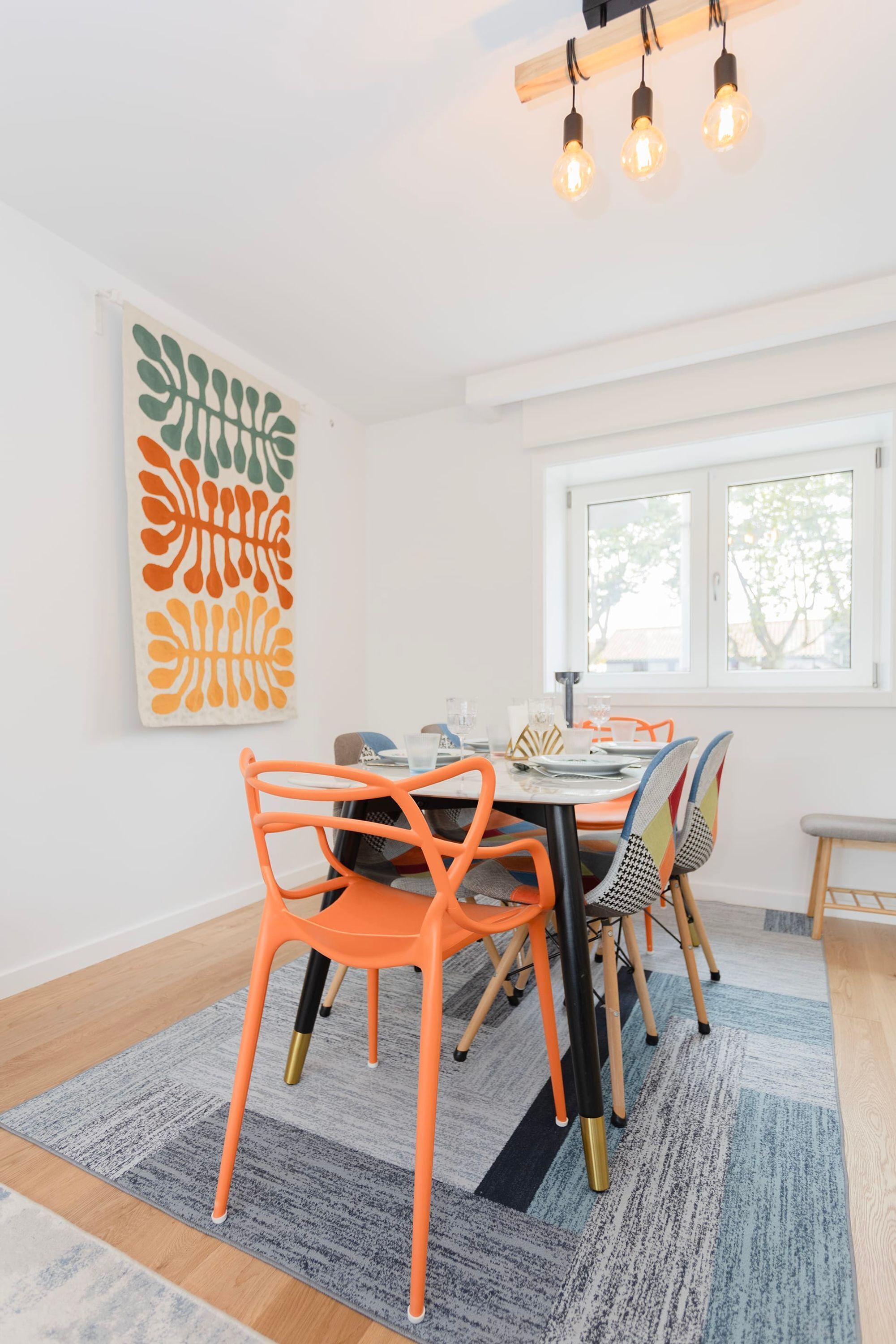 A dining area with a table set for four, surrounded by colorful chairs, a patterned rug, and a large abstract painting on the wall.