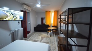 A hotel room with a bed, a small table, a chair, a TV mounted on the wall, a ceiling fan, and a window with curtains. - Mami Affittacamere (Genoa)