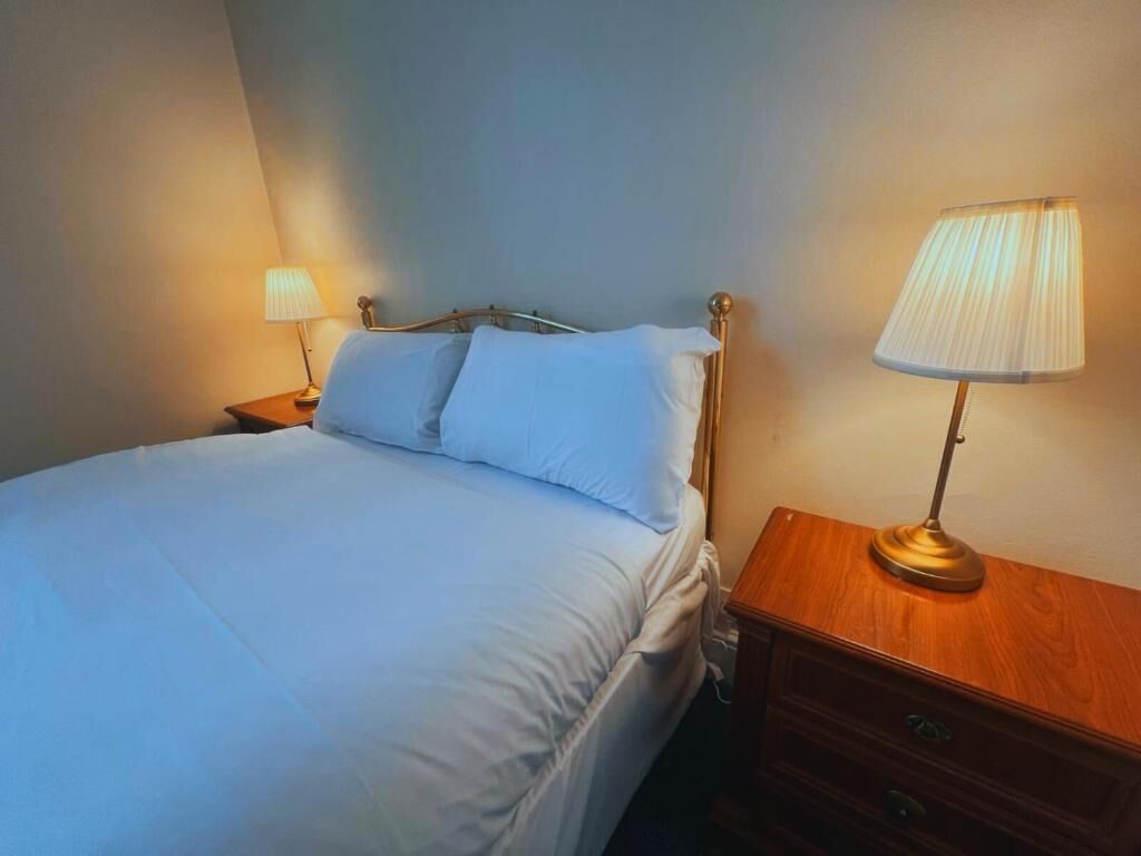 Una cama bien hecha con sábanas blancas, una mesita de noche de madera con una lámpara y un estante fijado a la pared.
