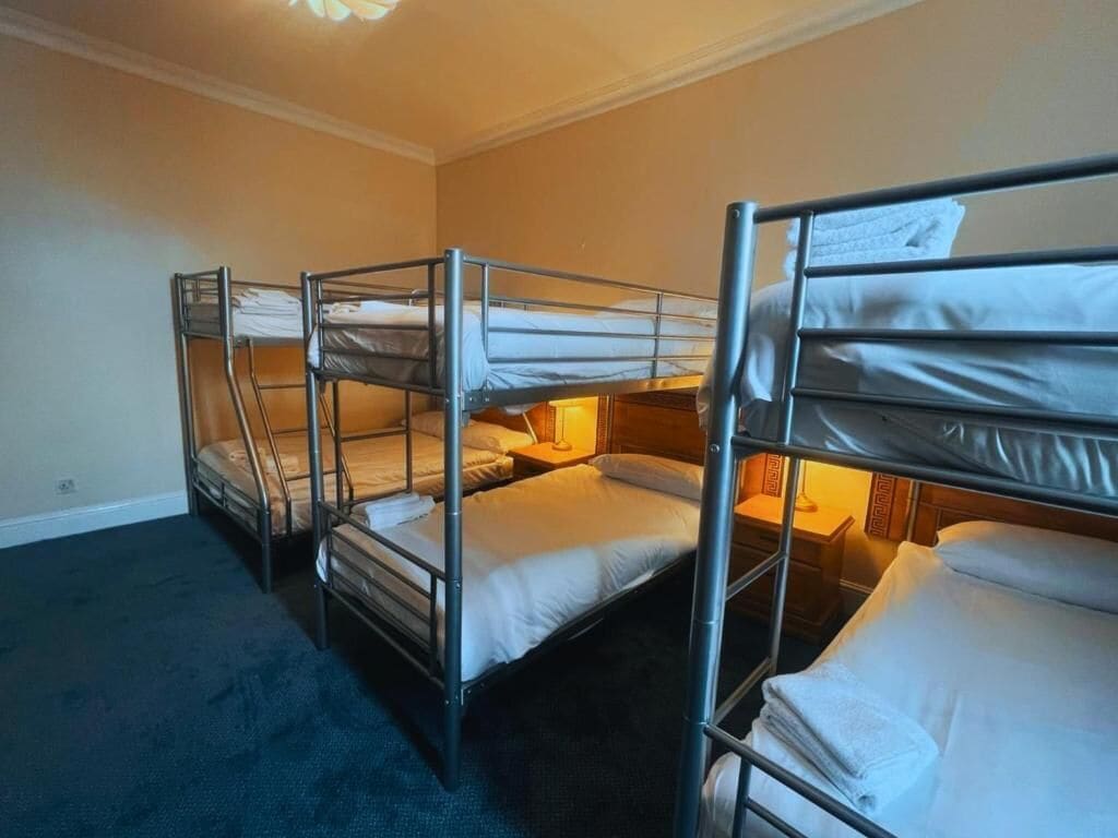 A room with bunk beds arranged in a row, a carpeted floor, and a bedside lamp.
