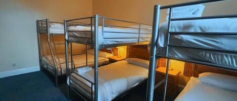 A room with bunk beds arranged in a row, a carpeted floor, and a bedside lamp.