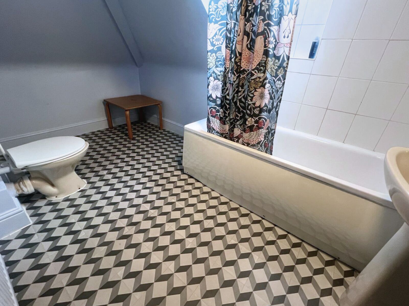 Ett badrum med ett schackrutigt golv, en toalett, en liten träpall och ett badkar med ett duschdraperi med blommönster.