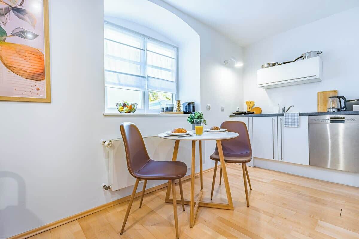 A modern kitchen with a dining area, featuring a large window, a stainless steel dishwasher, and a white cabinet.