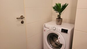 Una lavadora blanca y compacta en un lavadero pequeño, con una planta en maceta encima.