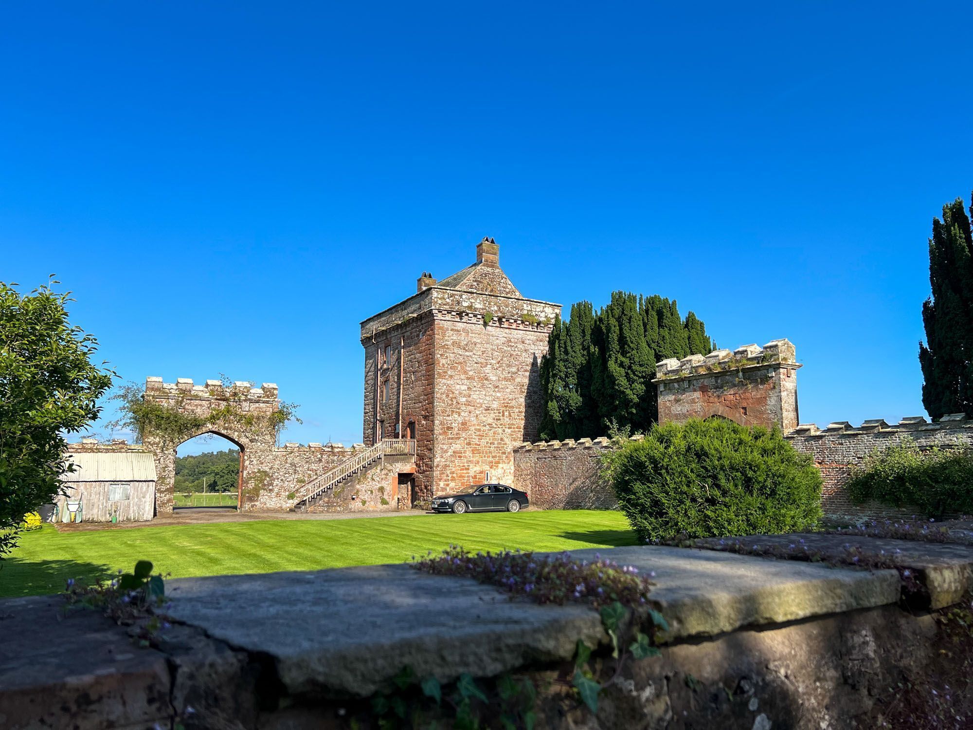 A historic stone building with a tower, surrounded by greenery and a clear blue sky.