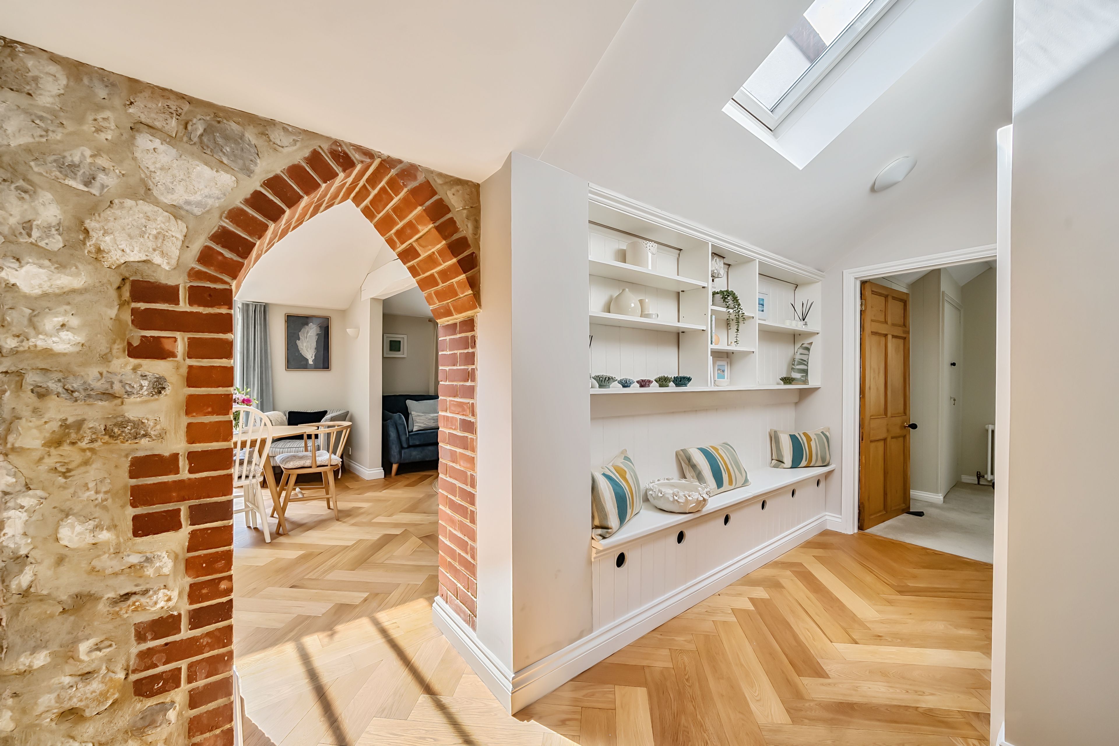 A spacious hallway with a herringbone wooden floor, a brick archway, and a white shelving unit with decorative items.