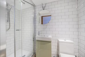 Et badeværelse med hvide flisevægge, en glasvægs brusekabine, en vask og et toilet.
