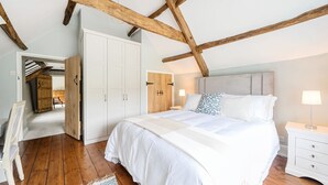A bedroom with a large bed, wooden beams, white wardrobe, and a nightstand with a lamp. - Dovehayes Cottage  - Bourton, Shaftesbury (Gillingham)