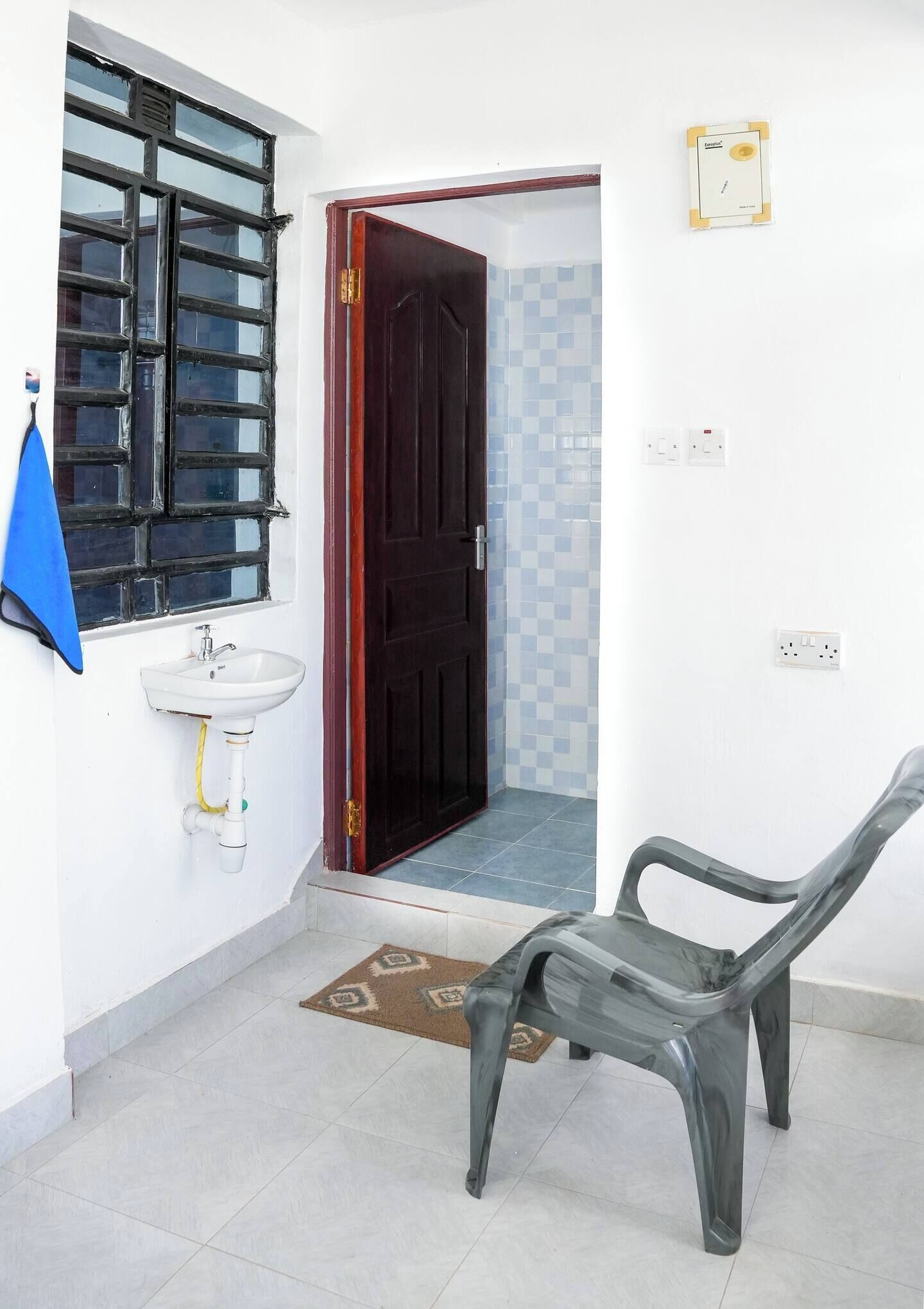 Una habitación con pared blanca, una ventana con persianas negras, una puerta de madera, un lavabo blanco y una silla de plástico.