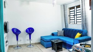 Una sala de estar con un sofá azul, dos taburetes azules, una mesita negra y un ventanal con vistas.