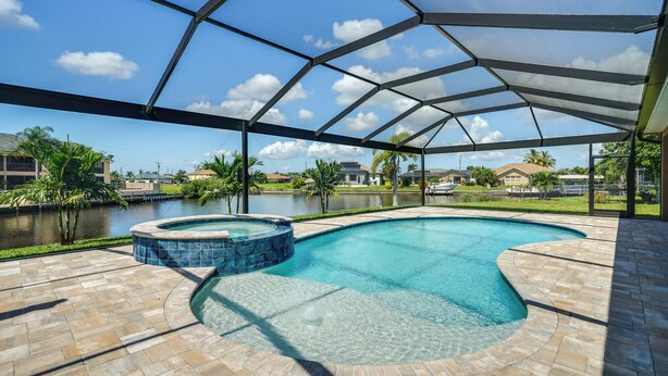 A swimming pool with a hot tub, surrounded by a covered patio area with a view of a lake and houses in the background. - Cape Coral Villa: Heated Pool & Gulf Access! (Cape Coral)